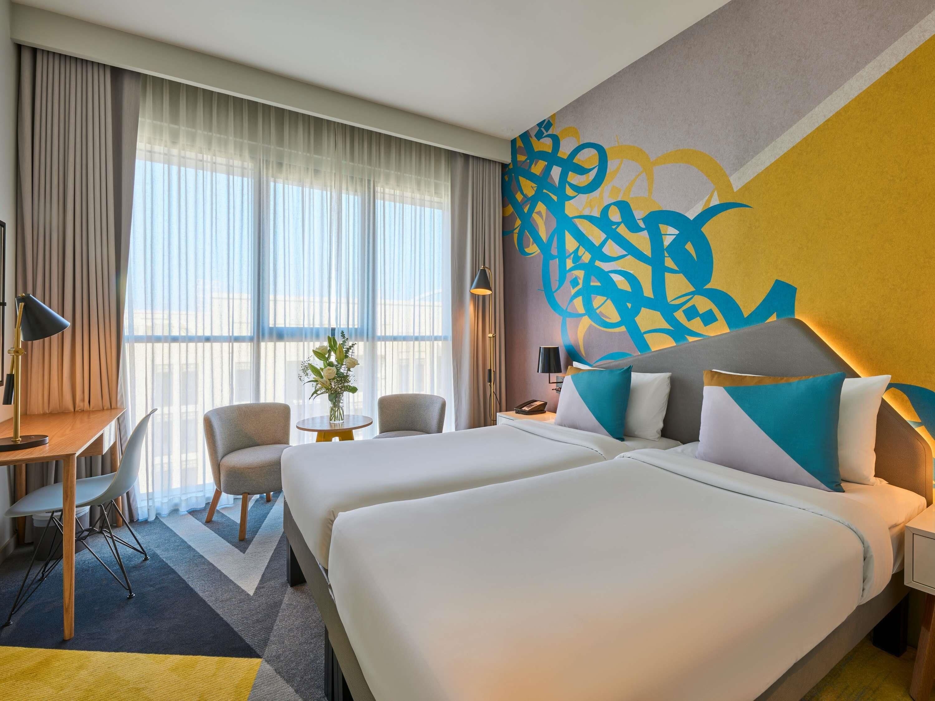 Hotelkamer met twee bedden, een bureau, een stoel, een kleine tafel met een bloevas en een groot abstract wanddecor.