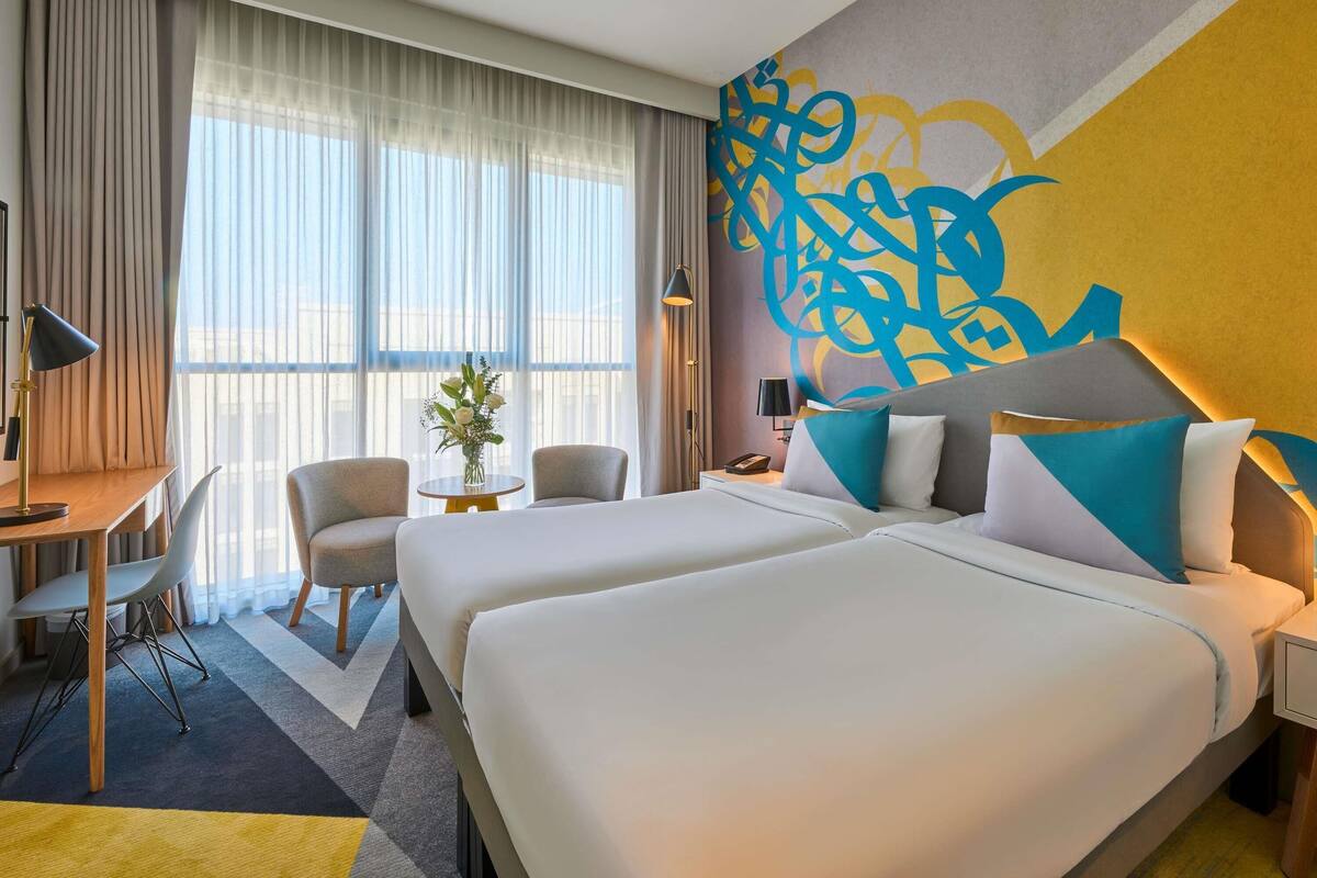 Ein Hotelzimmer mit zwei Betten, einem Schreibtisch, einem Stuhl, einem kleinen Tisch mit Blumen und einem großen abstrakten Wandbild.