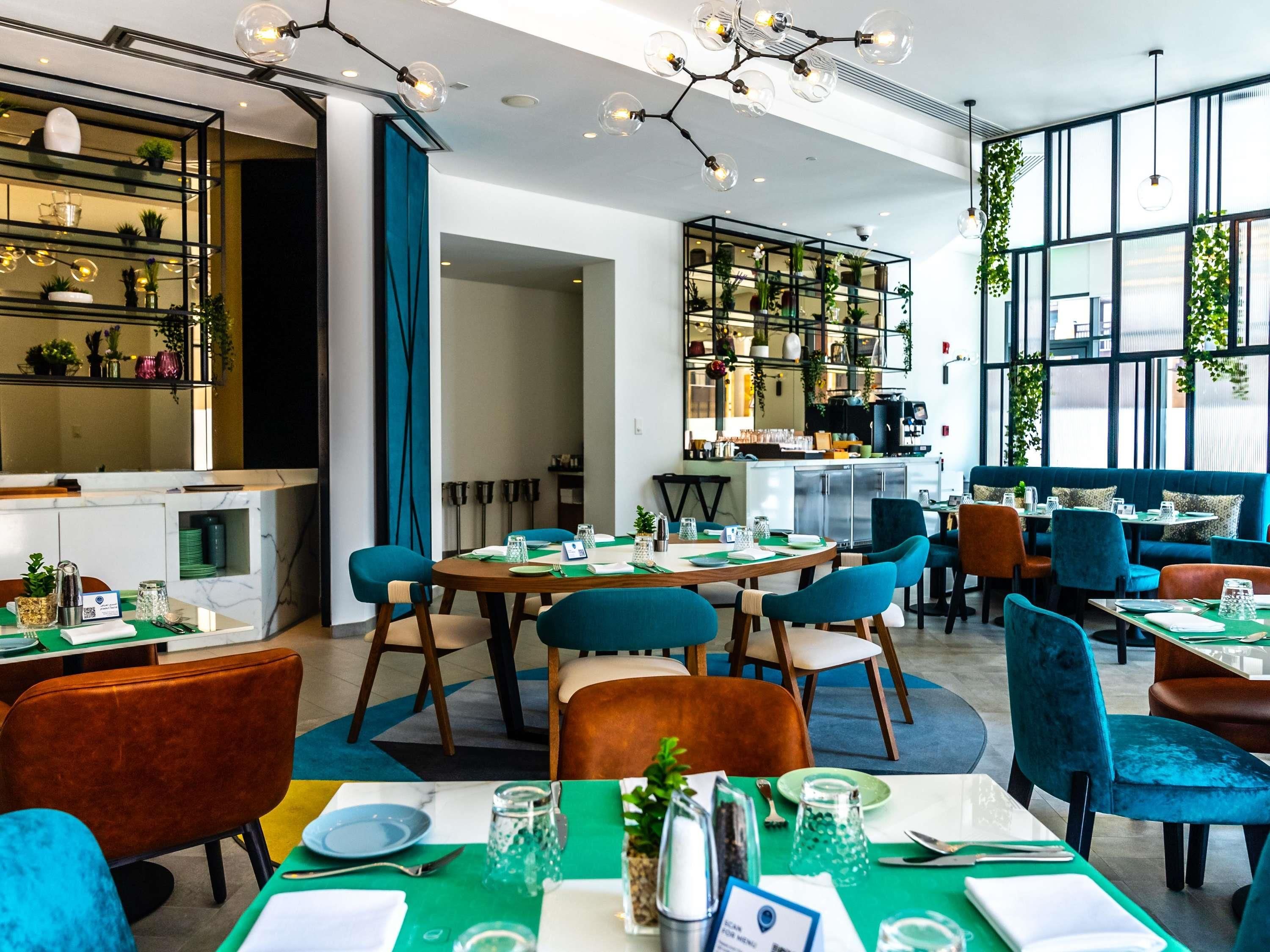 A modern restaurant with round tables, teal and brown chairs, and a kitchen area visible through a glass partition.