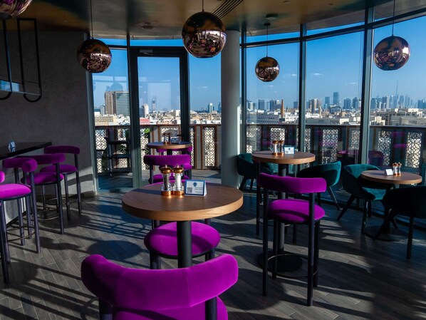 Un área de estar moderna en interiores, con mesas redondas y sillas moradas, amplias ventanas con vista a la ciudad.