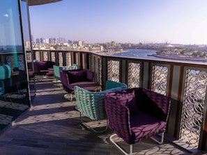 An outdoor seating area with cushioned chairs and tables, overlooking a cityscape with buildings and a waterfront. - Ibis Styles Dubai Deira (Dubai)