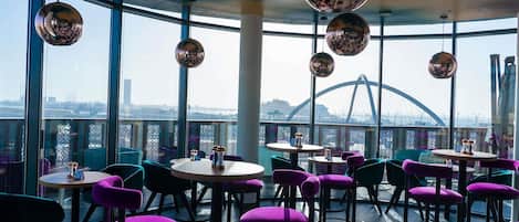 A rooftop restaurant with round tables and purple chairs, large windows, and a view of a bridge.
