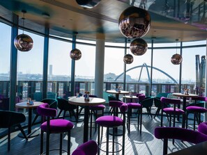 Un restaurant situé sur le toit d’un immeuble, avec des tables rondes et des chaises violettes, de grandes fenêtres et une vue sur un pont.