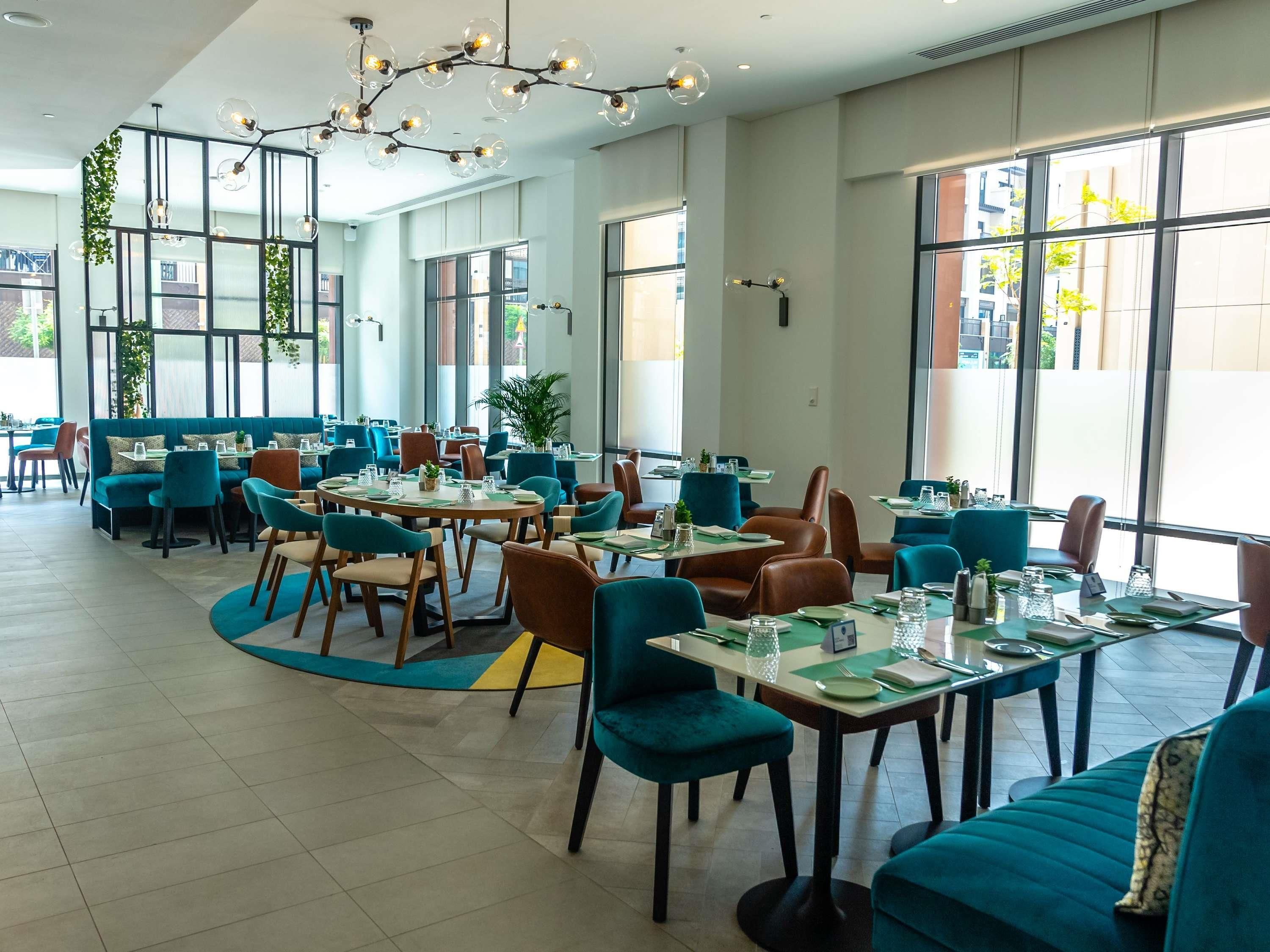 A modern dining area with round tables, teal and brown chairs, and large windows offering views of the city.