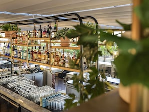 Un espace bar avec des étagères remplies de bouteilles d’alcool, de verres et une plante au premier plan.