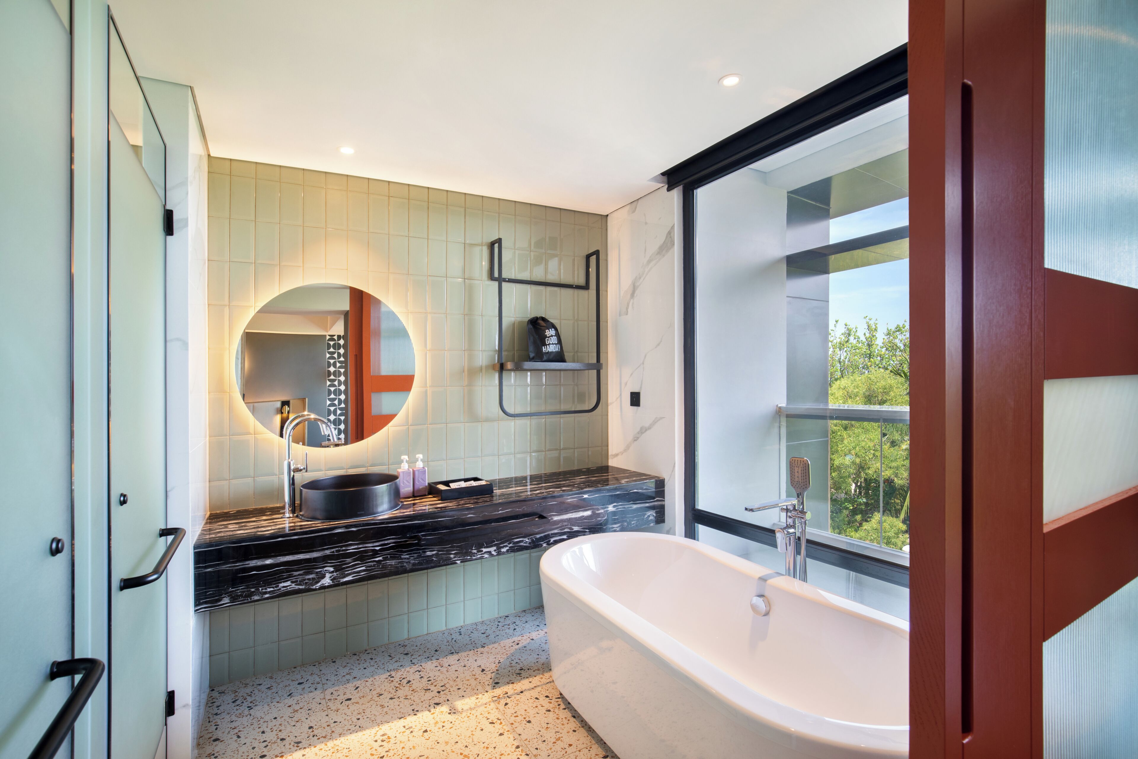 A modern bathroom with a freestanding bathtub, a large mirror, and a window providing a view of greenery.