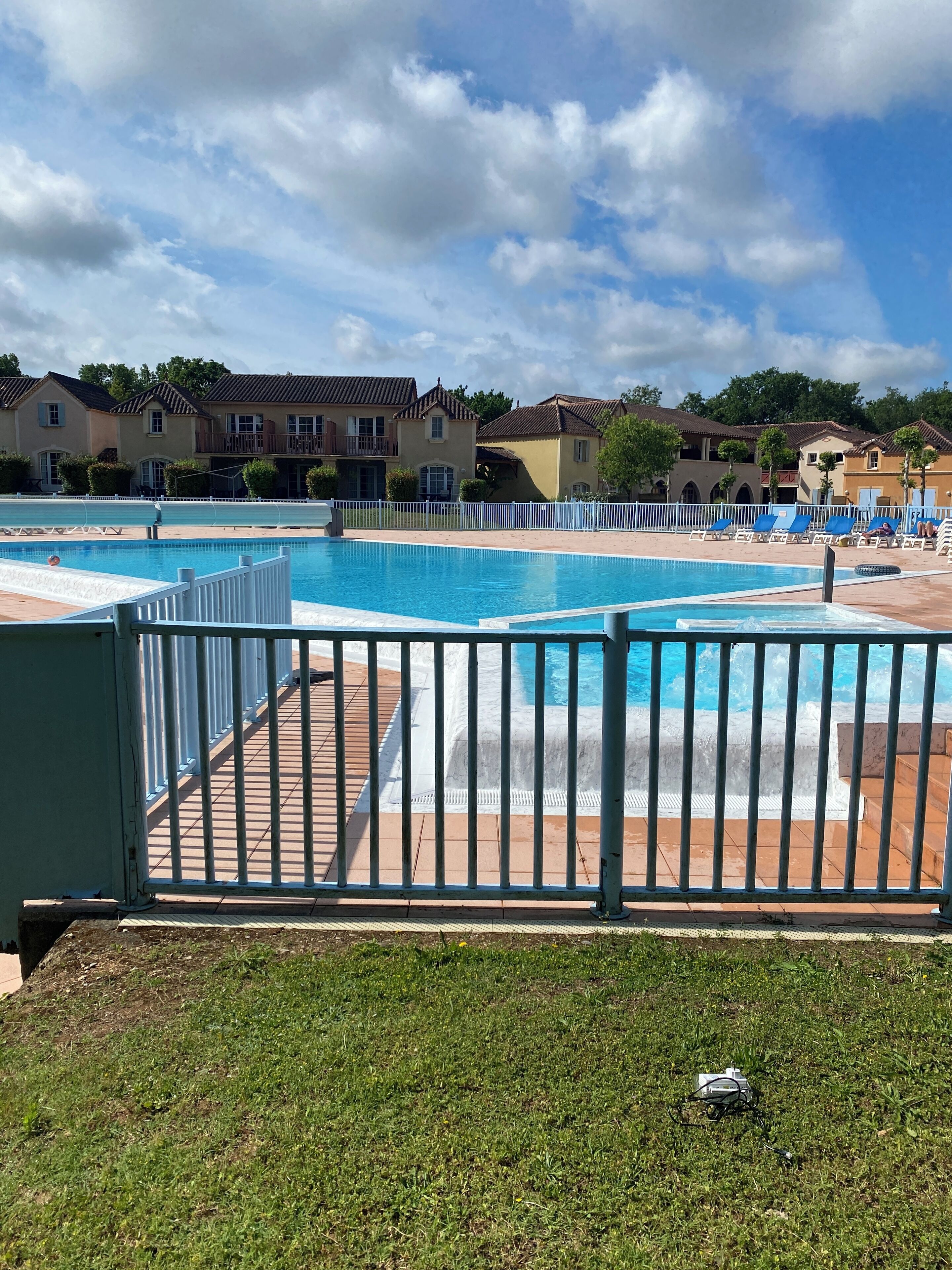 Una piscina con cerramiento blanco, rodeada de césped y algunos edificios al fondo.