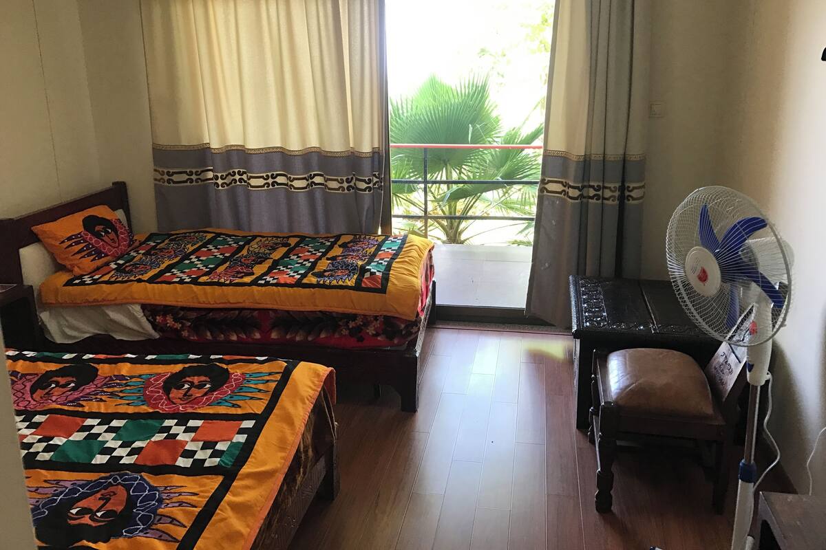 Un dormitorio con dos camas, un escritorio con silla, un ventilador de pie y una ventana con cortinas.