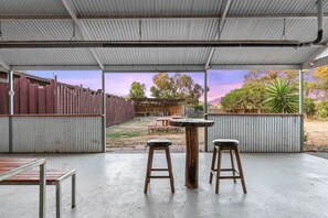 An outdoor patio area with a table and two stools, a wooden fence, and a view of trees and a building in the background. - Torrumbarry Motel (Torrumbarry)