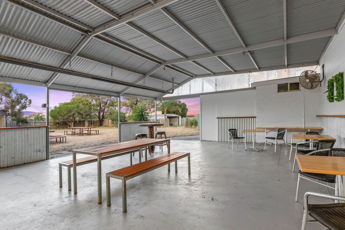 Un espacio al aire libre techado con mesas y bancos, una pared blanca con una ventana y vistas a árboles y al atardecer.