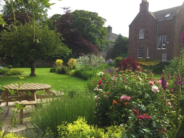 A garden with a wooden table and chairs, surrounded by various flowering plants and trees.