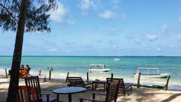 A beachside seating area with wooden chairs and tables, overlooking a calm sea with a boat anchored nearby. - Stay.Plus Bamburi Beachfront Apartment (Mombasa)