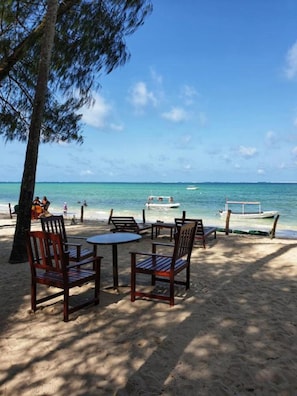 A beachside seating area with wooden chairs and tables, overlooking a calm sea with a boat anchored nearby.