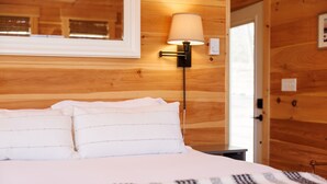 A wooden-paneled room with a bed, a nightstand, a lamp, and a door. - Quiet Maine Cabin 2 • Hideaway in the Trees • Sauna (Stockton Springs)
