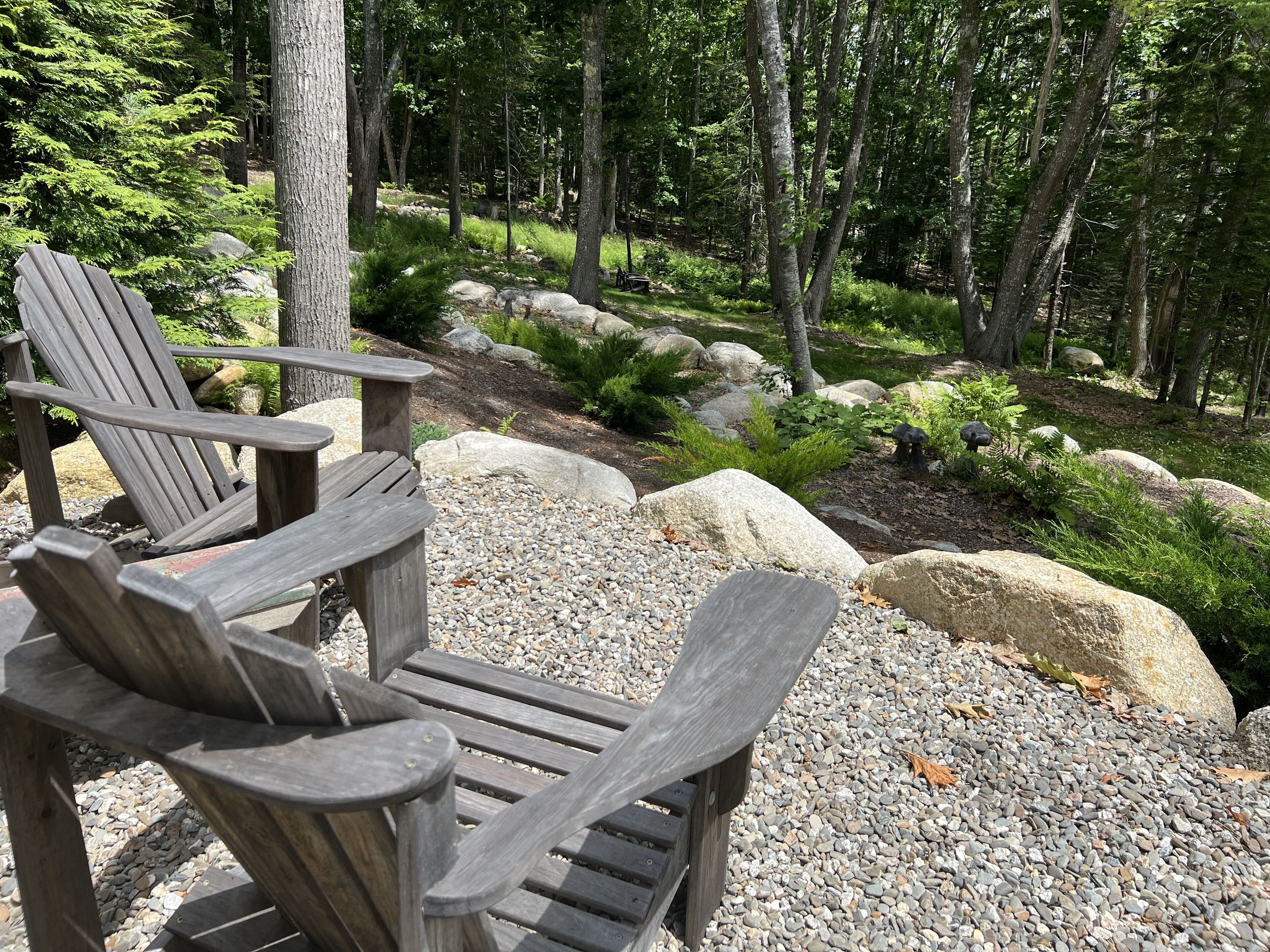 Eine Holzterrasse mit Adirondack-Stühlen und einem Kiesweg, umgeben von Bäumen und Steinen.