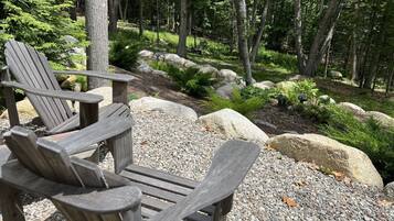 Eine Holzterrasse mit Adirondack-Stühlen und einem Kiesweg, umgeben von Bäumen und Steinen.