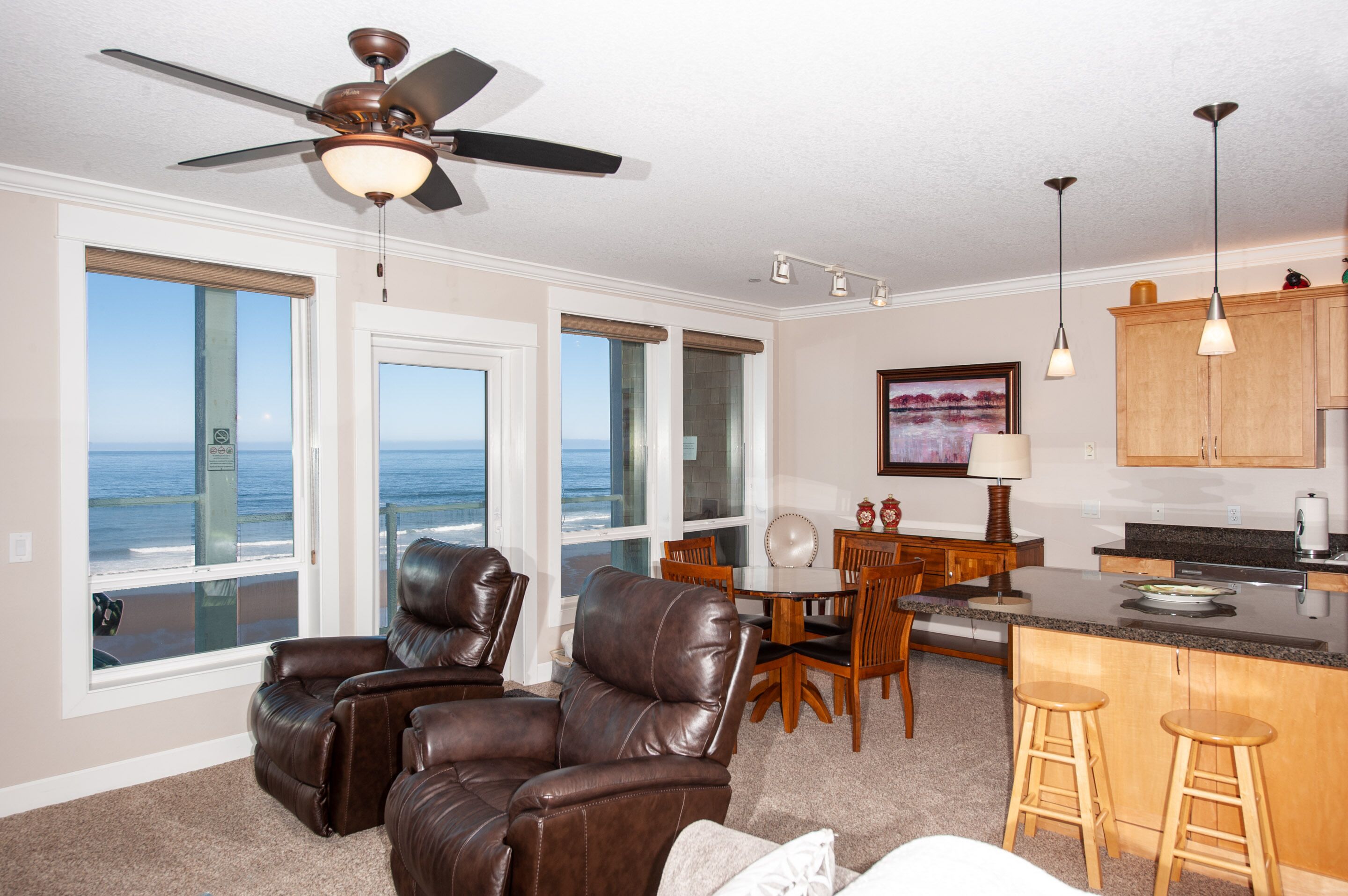 A spacious living area with a view of the ocean, a ceiling fan, a dining table with chairs, and a kitchen area with bar stools.