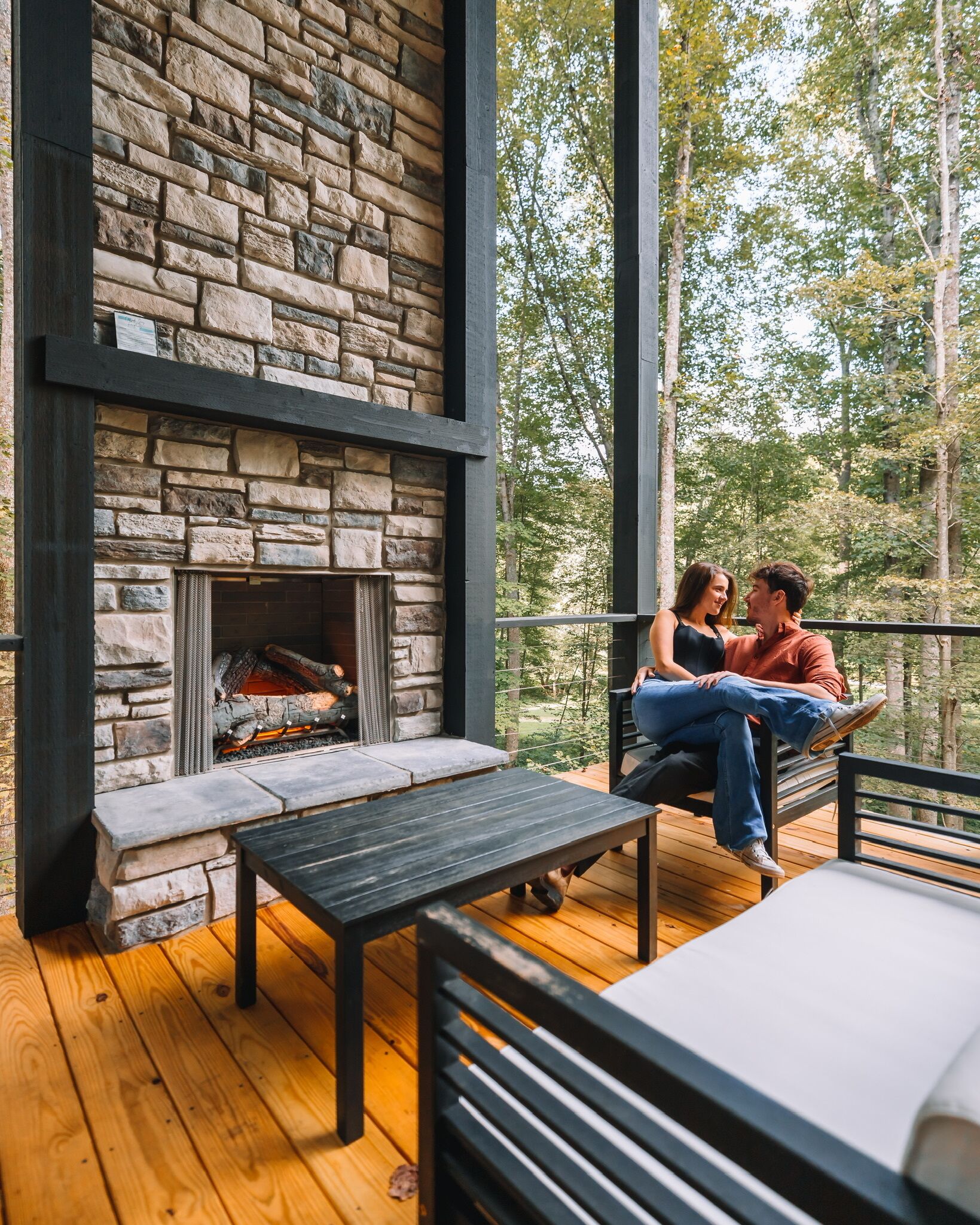 A stone fireplace with a bench and table in a wooden deck area, overlooking a forest.
