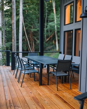 Une terrasse extérieure en bois, avec une table rectangulaire et des chaises, entourée de grands arbres.