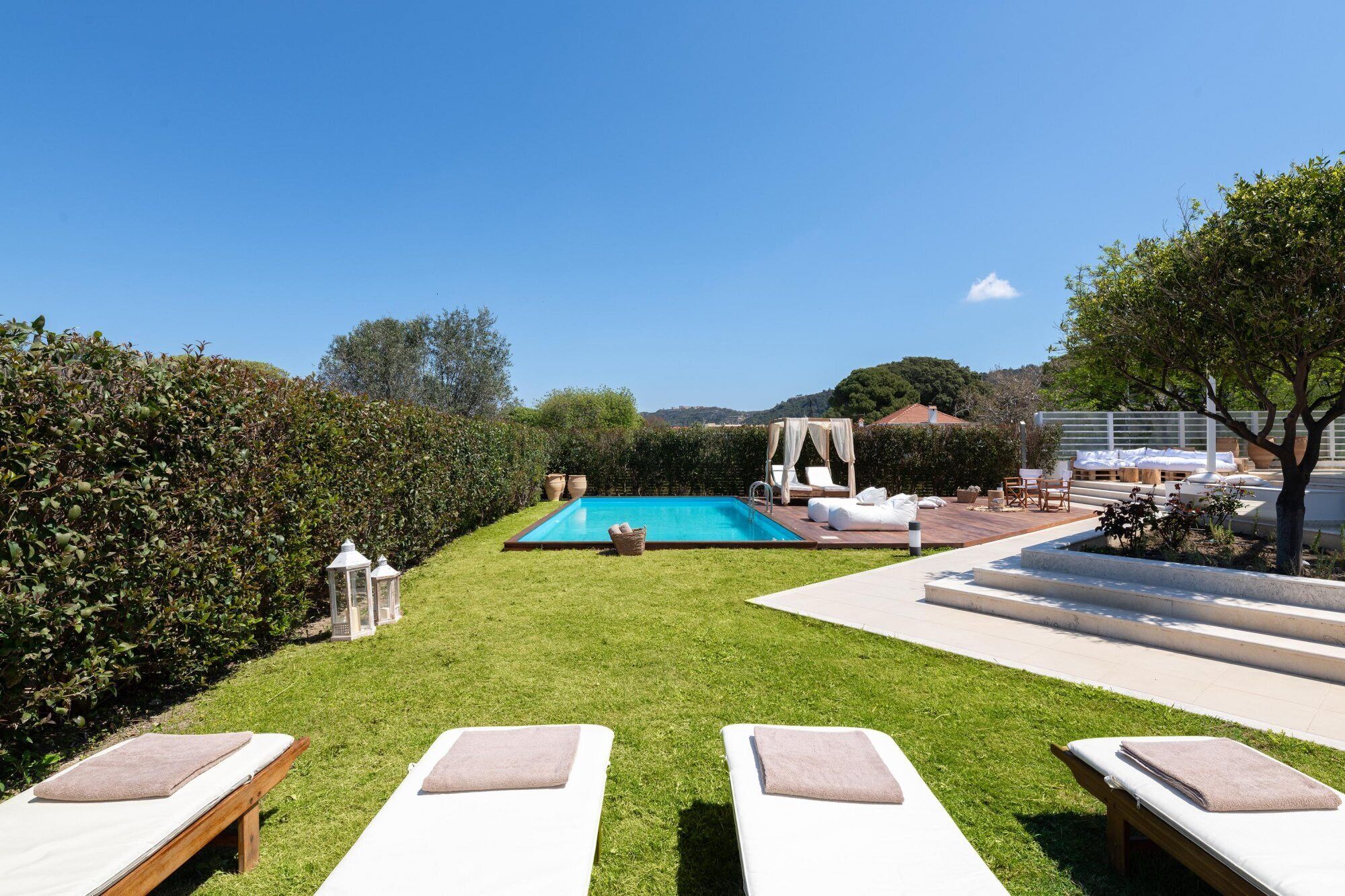 Zona de piscina con sillones de descanso y una pérgola, rodeada de césped y setos bien cuidados.