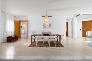 A spacious dining area with a wooden table and white chairs, a hanging chandelier, and a large window with curtains.