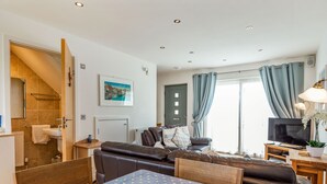 A modern living room with a sofa, a dining table with a polka dot tablecloth, a TV, and a staircase leading to a bathroom. - Constantine Bay Cottage (Padstow)