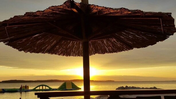 Um guarda-sol na praia com vista para o pôr do sol, mar calmo e ilhas ao longe.