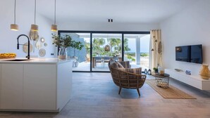 A modern kitchen with a central island, a dining area with a wicker chair, a TV, and a large window with a view of palm trees. - Blue Bay | The Reef | Terrace (Sint Michiel)