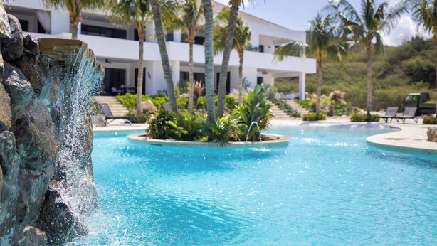 A swimming pool with a waterfall feature, surrounded by palm trees and a modern building. - Blue Bay | The Reef | Terrace (Sint Michiel)