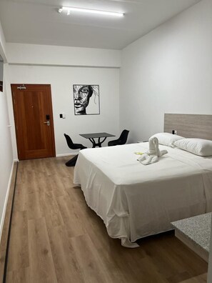 Quarto de hotel com cama, uma mesinha, duas cadeiras, porta de madeira e uma obra de arte na parede.