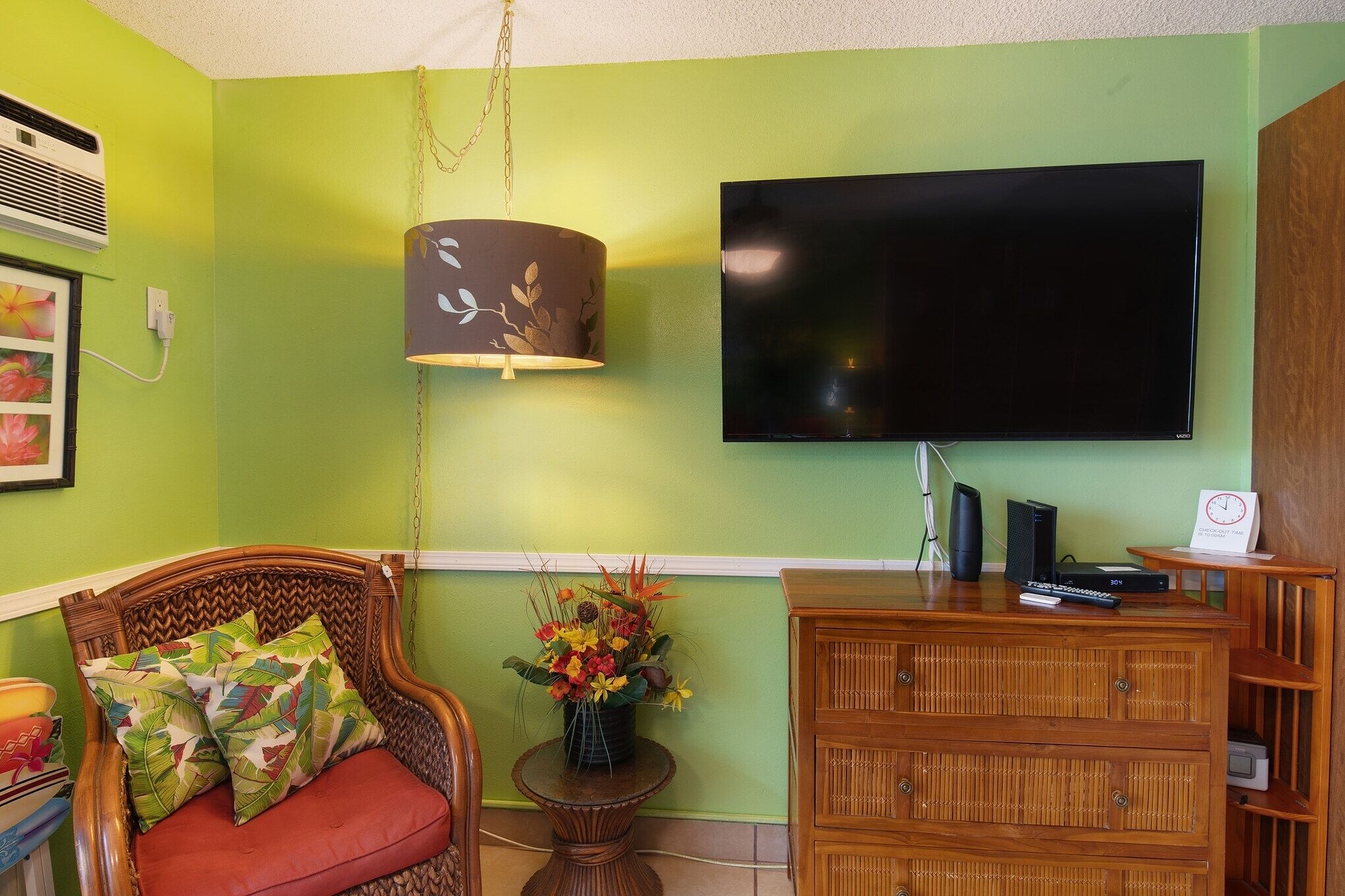 A living room with a green wall, a flat-screen TV, a wicker chair with red cushions, a small table with a vase of flowers, and a wooden cabinet.