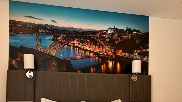 A hotel room with a large bed, two bedside tables, a nightstand, a wall-mounted lamp, a telephone, and a view of a cityscape with a bridge and buildings. - Hotel Porto Interface Trindade By Kavia (Porto)