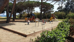 A playground with a slide and climbing structures, surrounded by trees and a sandy area. - At the end of the garden, the pine forest and the beach (Poggio-Mezzana)