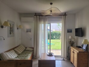 Un salon comprenant un canapé, une table, un téléviseur et donnant sur un jardin grâce à une porte coulissante.