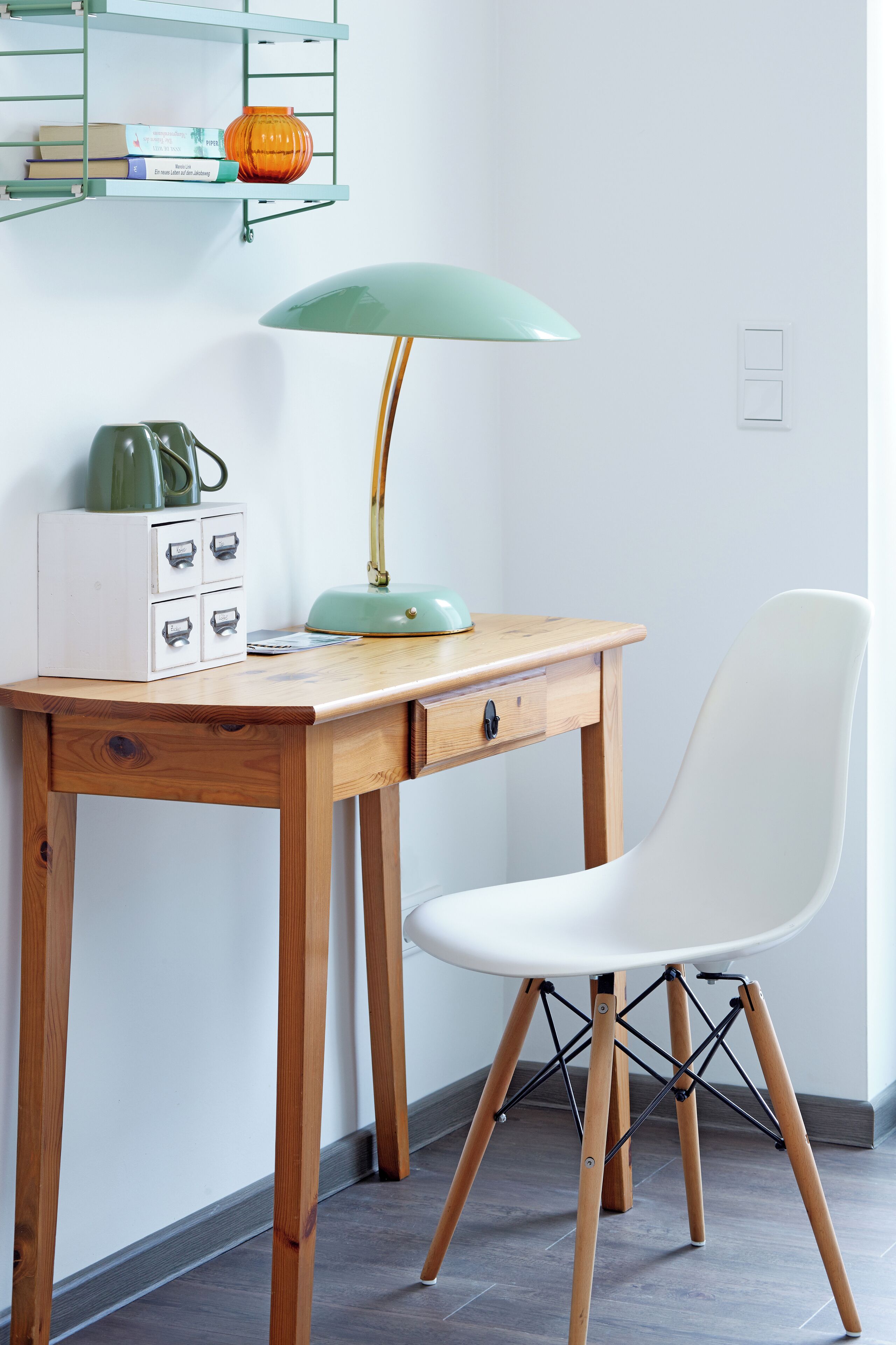 Un bureau en bois, une chaise blanche, une lampe verte et un meuble de rangement blanc avec des tiroirs.