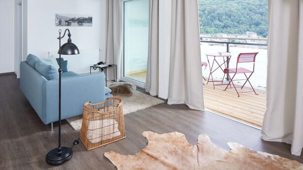 A modern living room with a sofa, a standing lamp, a wicker basket, and a large window with a view of a balcony and forested hills.