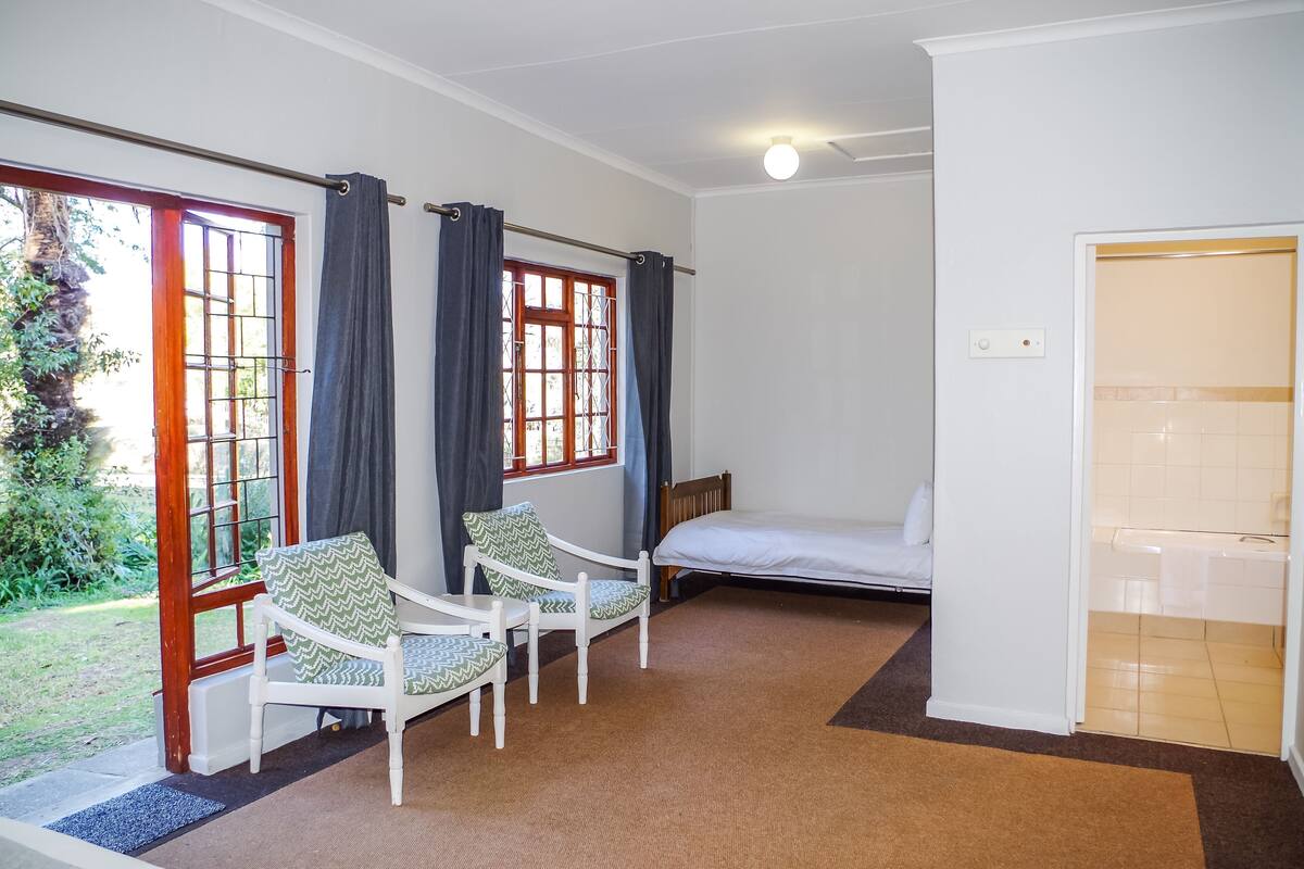 Une pièce comprenant un lit, deux chaises, une fenêtre avec des rideaux et une porte donnant sur une salle de bain.