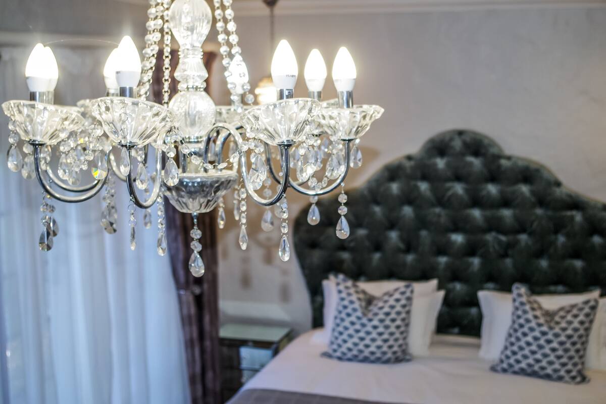 Un lustre à plusieurs lampes, orné de décorations en cristal, dans une chambre à coucher dotée d’une tête de lit capitonnée et de coussins à motifs.