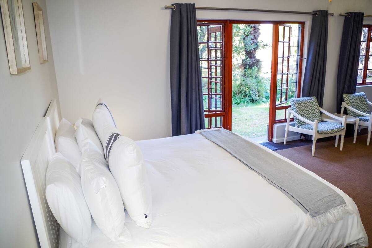 Une chambre avec un grand lit, une vue sur le jardin grâce à une porte vitrée, et deux fauteuils près de la fenêtre.