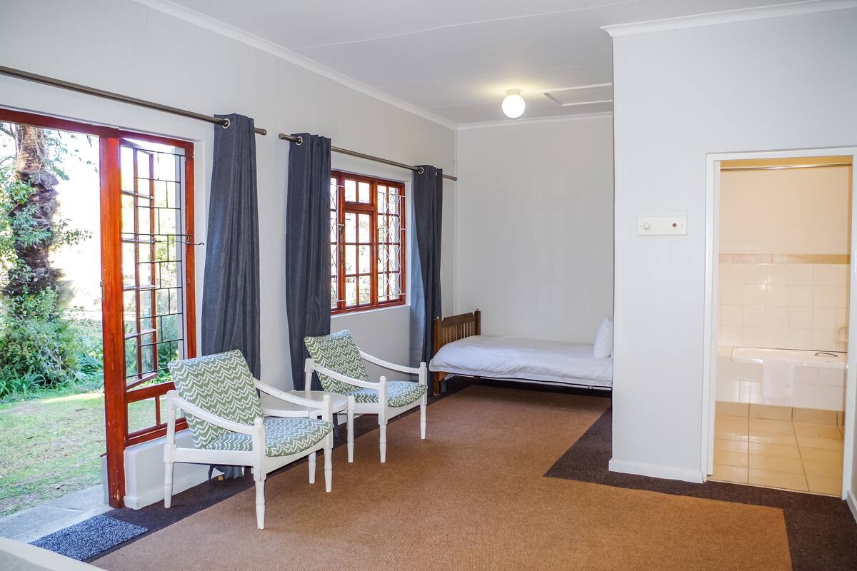 Une pièce comprenant un lit, deux chaises, une fenêtre avec des rideaux et une porte donnant sur une salle de bain.