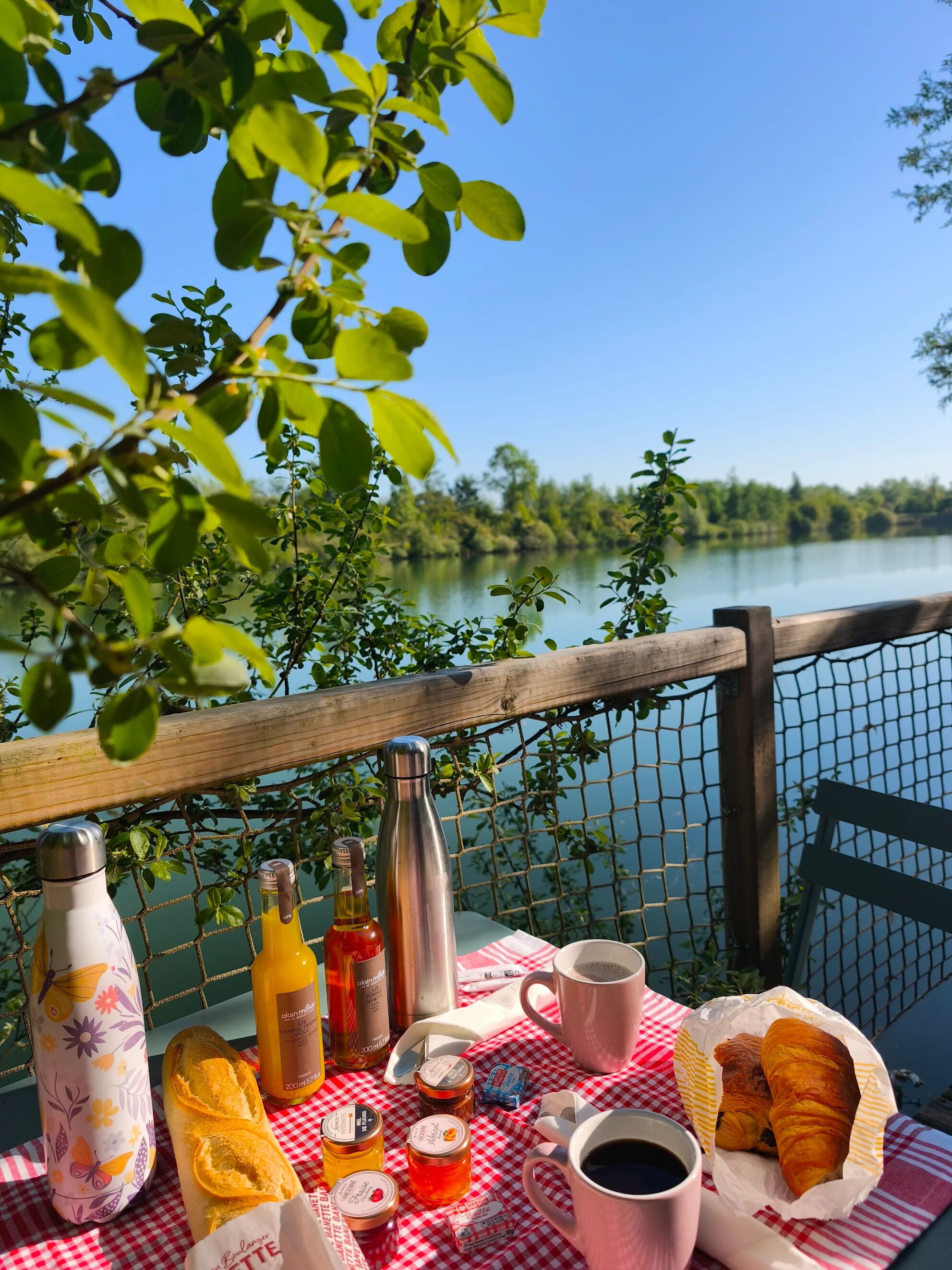 A picnic setup with a bottle of orange juice, a thermos, a bottle of wine, a jar of jam, a baguette, a croissant, and a cup of coffee on a checkered tablecloth overlooking a lake.