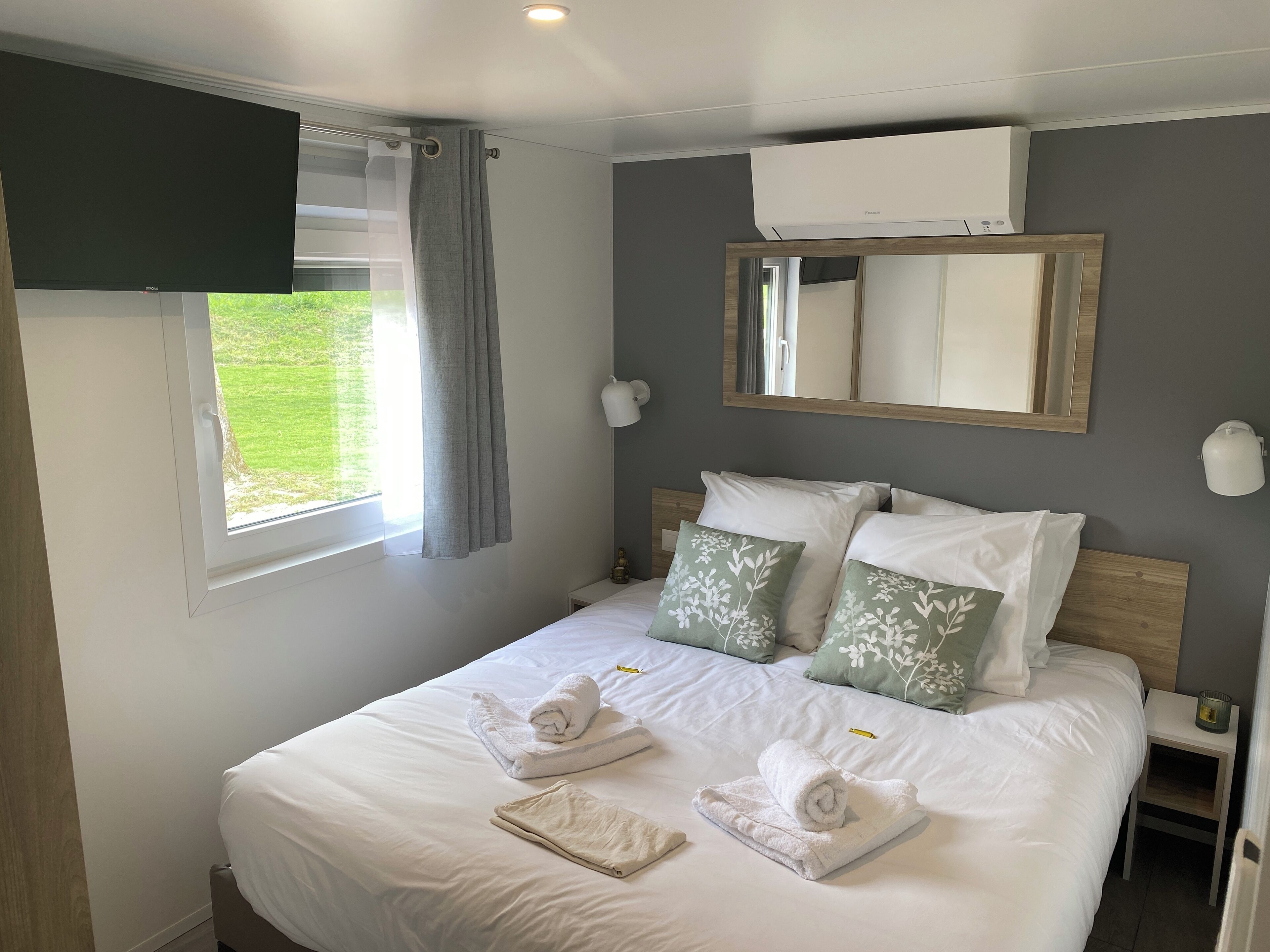 Un lit bien fait, avec du linge de lit blanc et des coussins à motifs verts, une table de chevet avec une bougie et une fenêtre avec des rideaux.