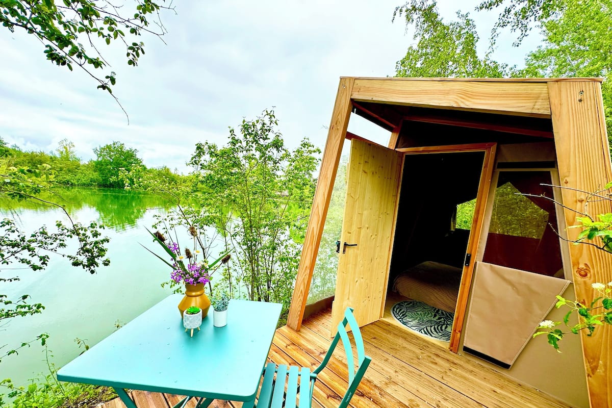 Una terraza de madera con una mesa y sillas, con vista a un lago tranquilo y abundante vegetación.