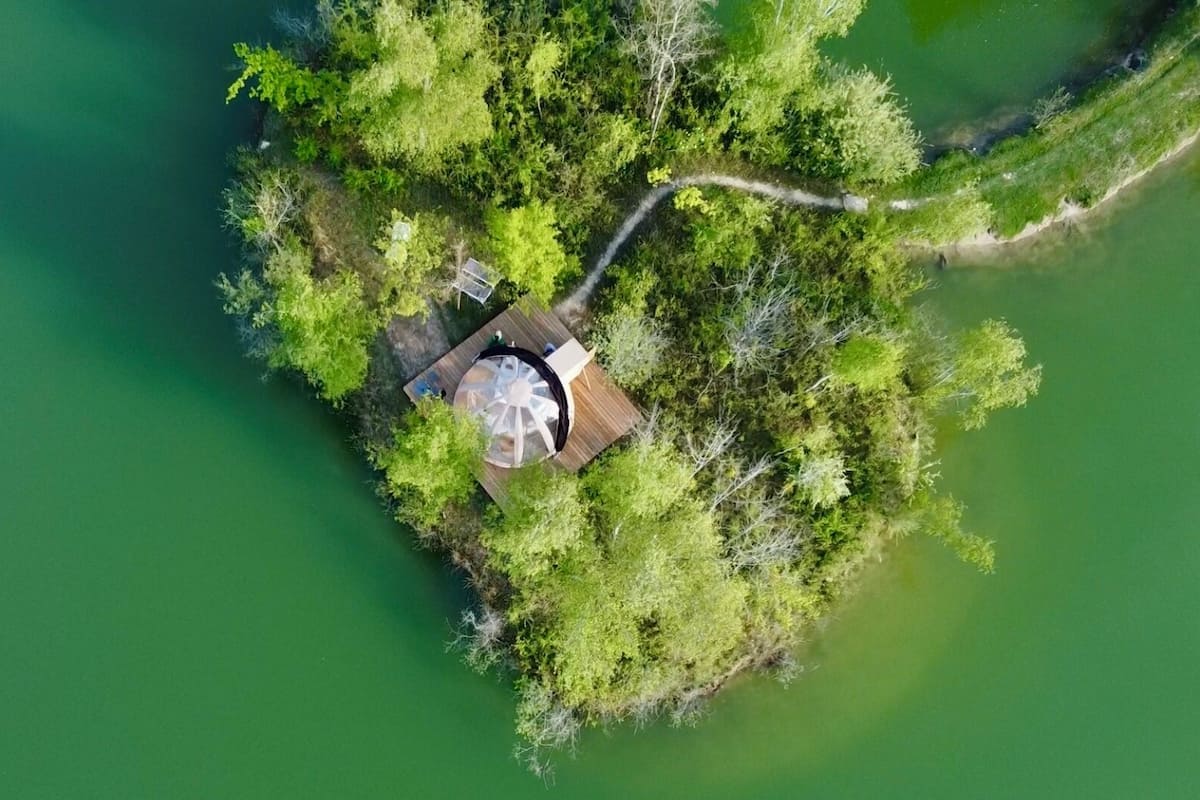 Vista aérea de una pequeña isla con un pabellón y una terraza de madera, rodeada de abundante vegetación y una masa de agua.
