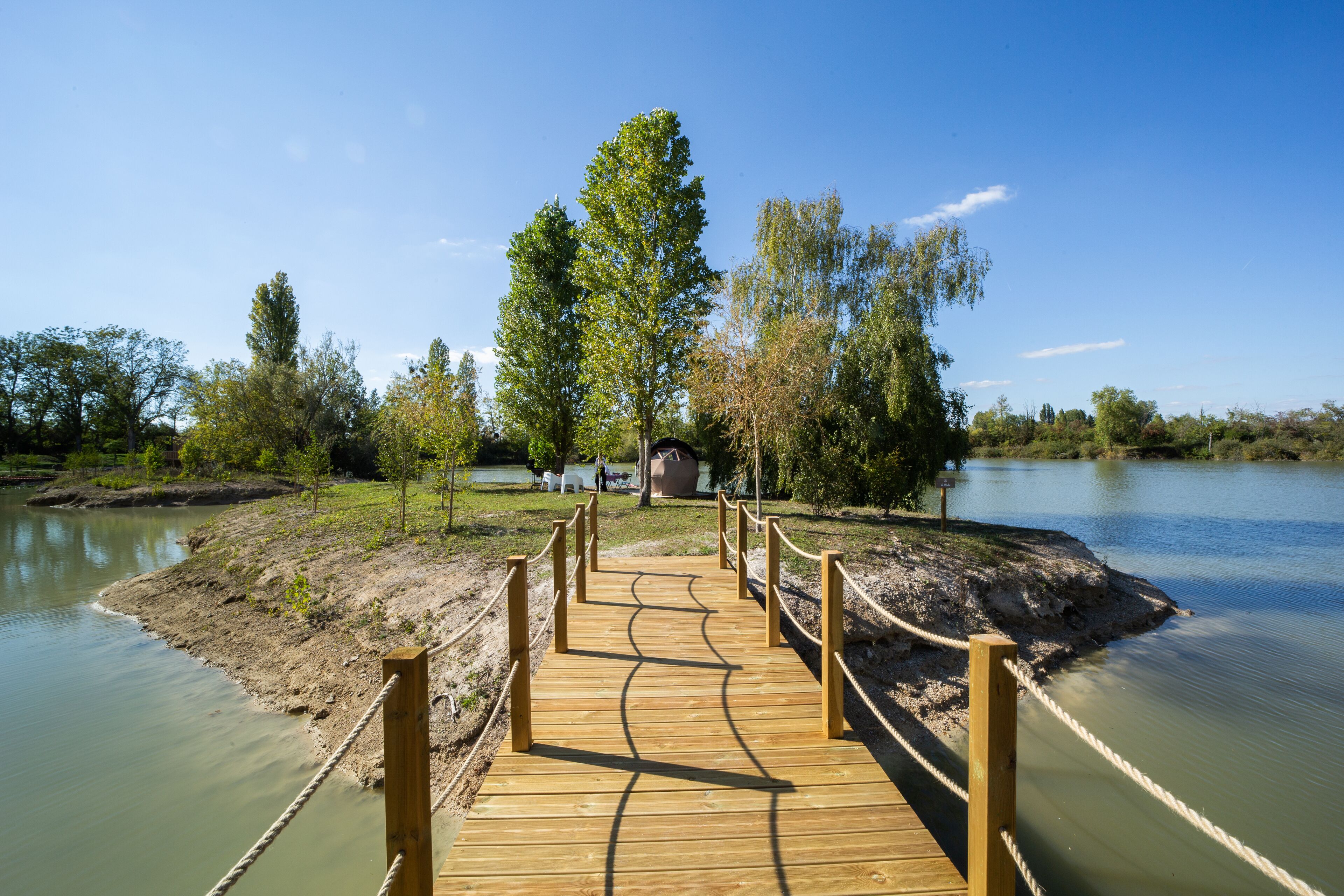 Un muelle de madera que lleva a una pequeña isla con un árbol grande y un edificio al fondo.