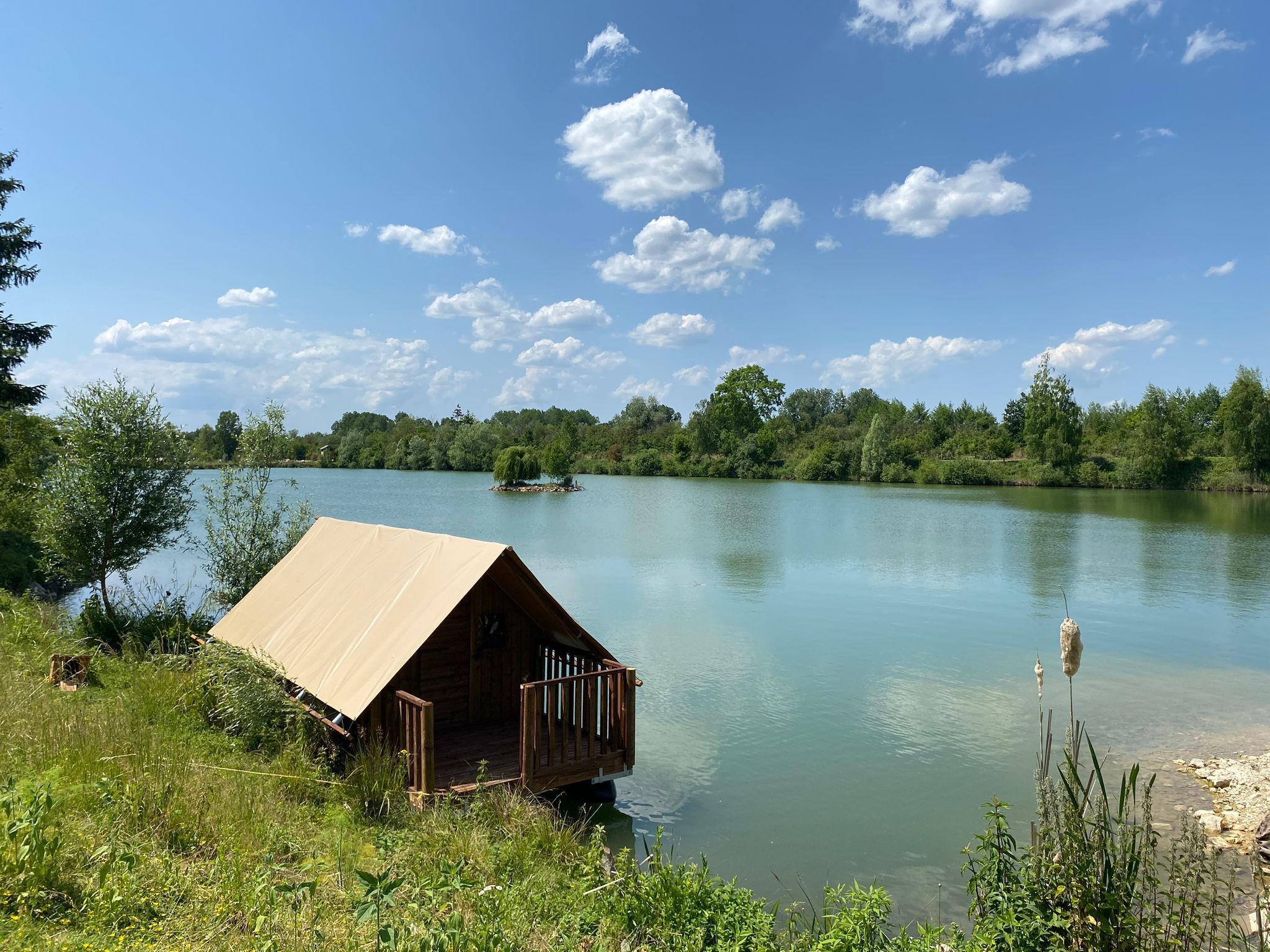 Una cabaña de madera con techo de metal junto a un lago tranquilo, rodeada de exuberante vegetación y árboles.