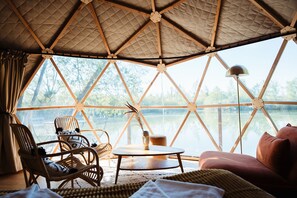 A cozy interior with a large window overlooking a lake, a round table, wicker furniture, and a cushioned seating area. -  Les Etangs de la Bassée (Gravon)