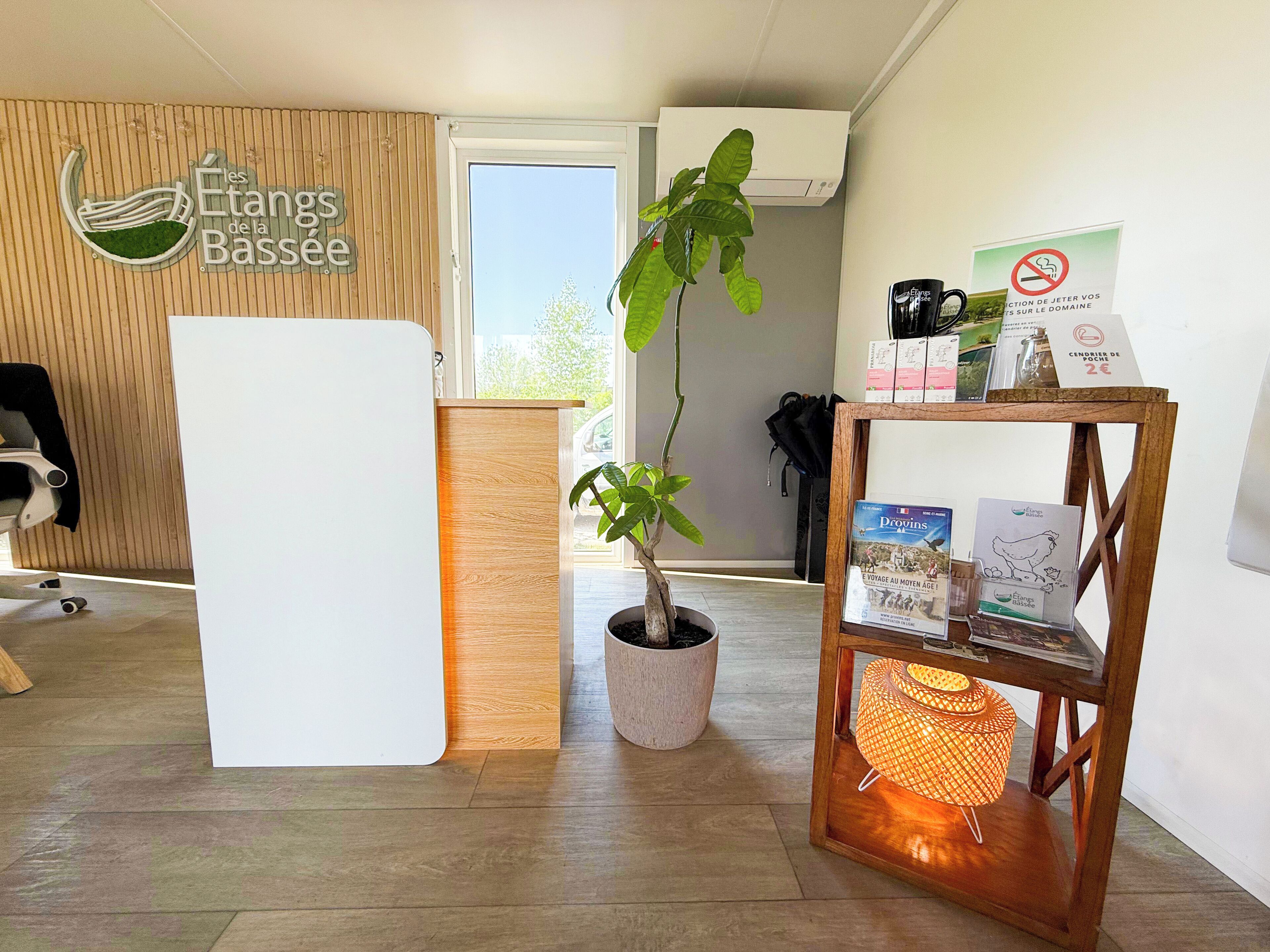 A reception area with a wooden reception desk, a potted plant, a wooden shelf with books and a candle, and a sign with a no-smoking symbol.
