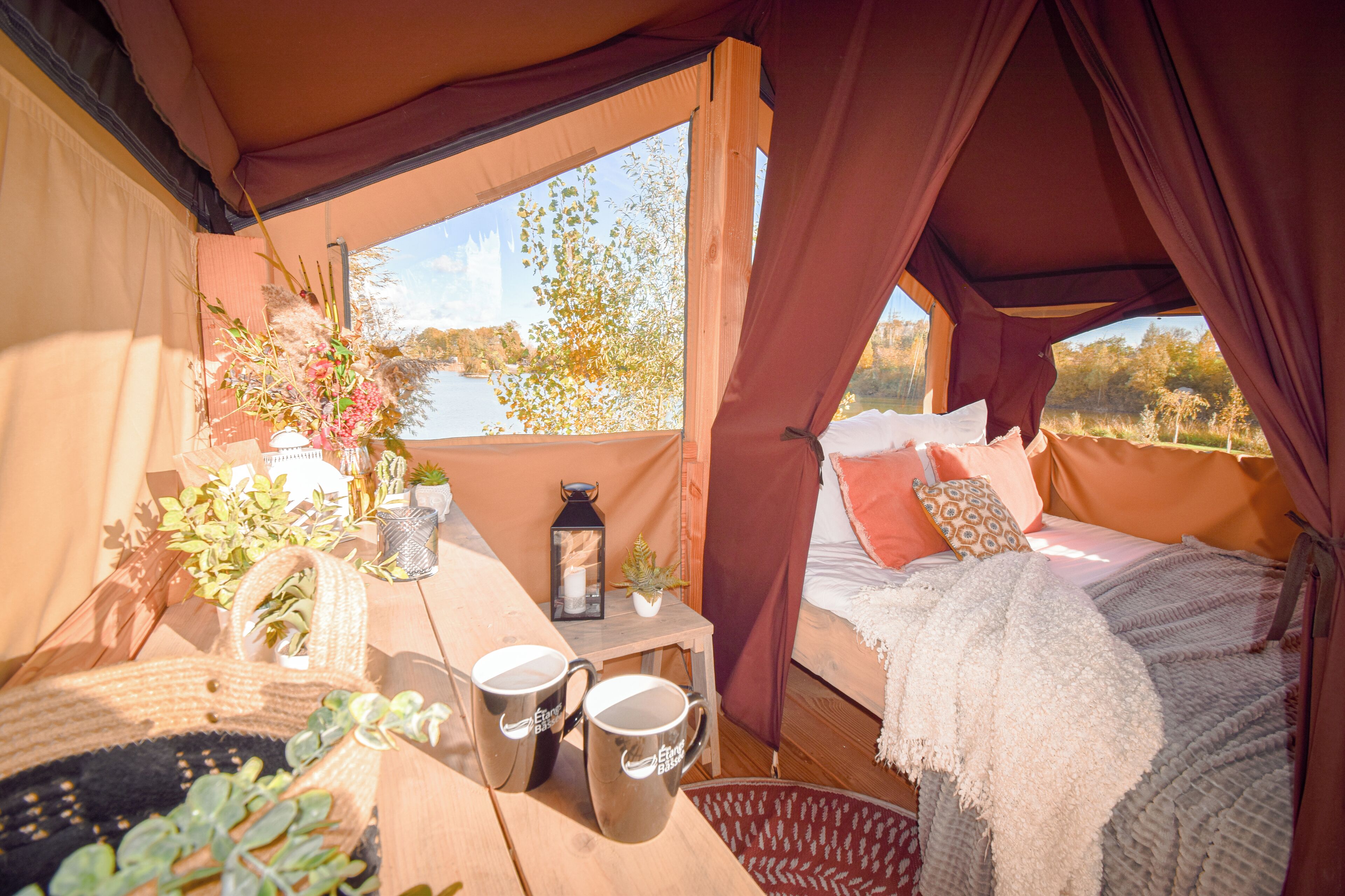 A cozy tent with a bed, a table with mugs, and a view of a lake.