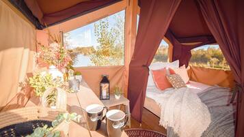 A cozy tent with a bed, a table with mugs, and a view of a lake.